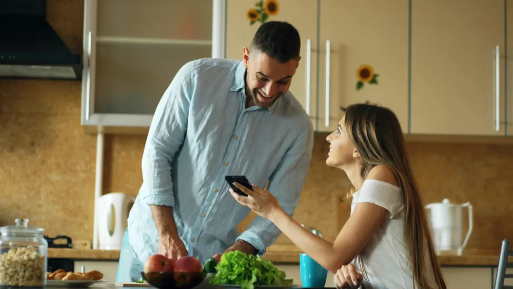 A couple in their kitchen smiling as they check something on a phone together