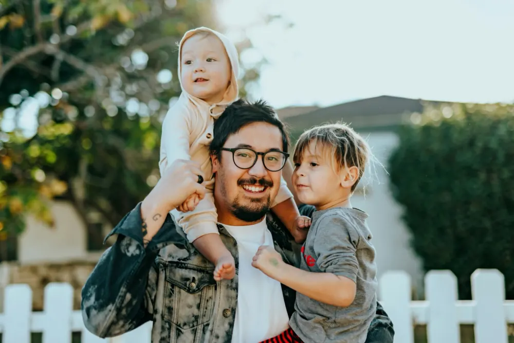 A smiling father outdoors carrying his two young children