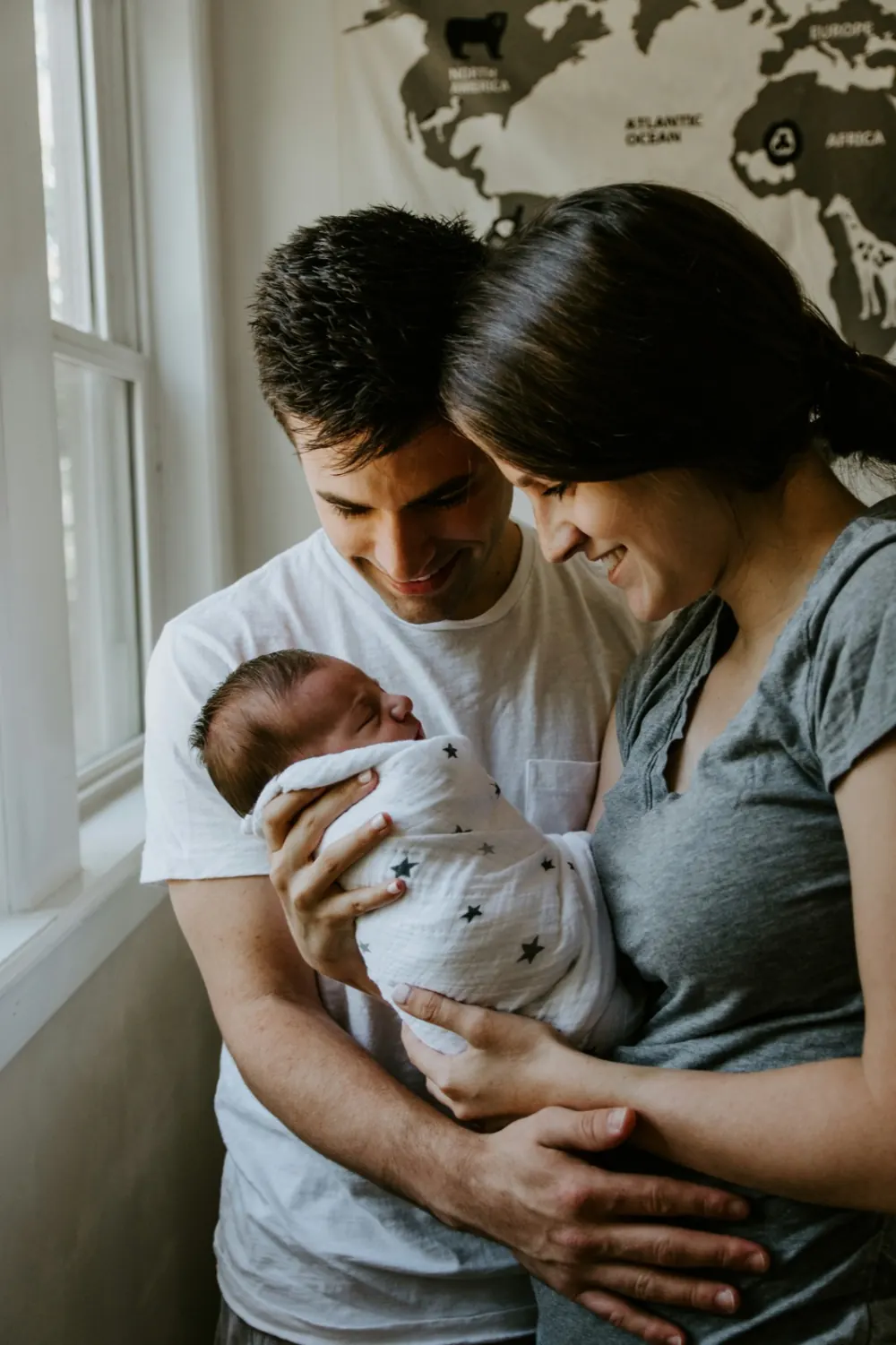 New parents holding their newborn baby by a window, a world map on the wall behind them